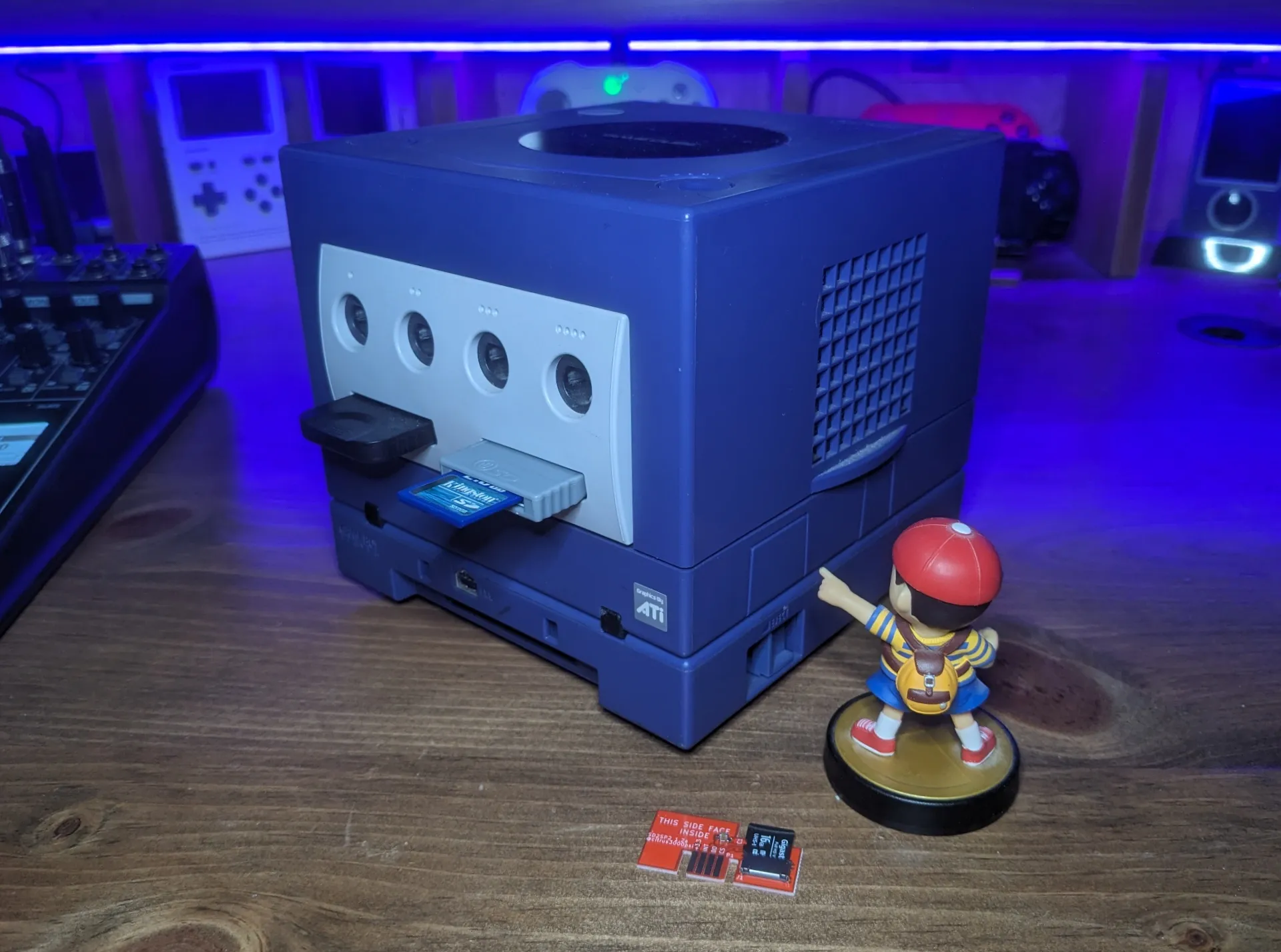 A Ness Amiibo points to the SP2 port on a GameCube with an sd2sp2 card laying on the desk near his pedestal.