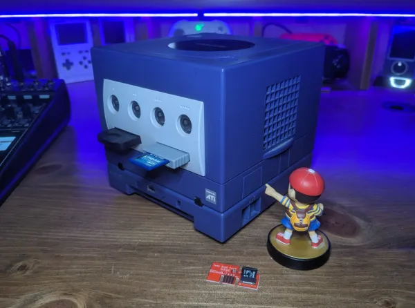 A Ness Amiibo points to the SP2 port on a GameCube with an sd2sp2 card laying on the desk near his pedestal.
